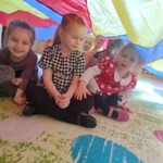 uśmiechnięte dziewczynki siedzą pod kolorową chustą animacyjną.