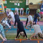 Dzieci stoją na dywanie i robią skłony_ trzymając pomarańczowe paski bibuły.