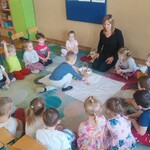 pani siedzi na krzesełku_ dzieci wokół. Chłopiec wkłada jajko do święconki.