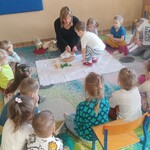 grupa dzieci siedzi  wokół białego materiału. Na środku pani i chłopiec wkłada coś do koszyka.