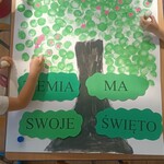 dłonie dzieci przyklejają różowe kwiatki do namalowanego drzewa_ niżej napis Ziemia ma swoje święto.