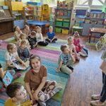 Dzieci siedzą na dywanie_ a przed nimi stoi dziewczynka z książkami w rękach.