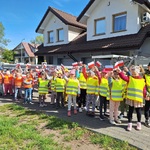 Dzieci w odblaskowych chorągiewkach stoją parami na chodniku przy domach_ trzymają w rękach chorągiewki.