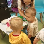 Dwójka dzieci stoi przy stoliku, za stolikiem stoi nauczycielka i robi tatuaż