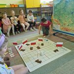 Dzieci siedzą wokół mapy Polski oraz maty_ na której stoją flagi.