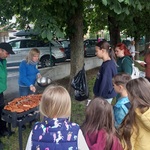 Na środku stoi grill z kiełbaskami_ Za grillem stoi pan_ Przygotowuje kiełbaski_ Rodzice i dzieci czekają na kiełbaski