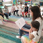 Pani czyta książkę_ obok niej siedzi jej córka_ w oddali dzieci słuchają bajki.