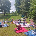 Dzieci siedzą na kocach pikniowych na placu przedszkolnym, machają w stronę osoby robiącej zdjęcia 