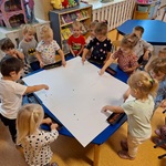 Grupa dzieci siedzi wokół stołu z dużym arkuszem papieru_ siedmioro dzieci stempluje farbami.