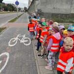 grupa dzieci w kamiselkach bepieczeństwa_ stoi na chodniku przy poziomych znakach ścieżki rowerowej.
