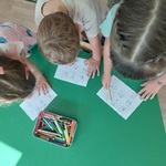 Troje dzieci pochylone nad stolikiem uzupełniają grę Bingo.