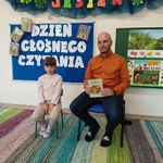 Dziewczynka siedzi obok taty trzymającego książkę na tle napisu Dzień Głośnego Czytania.