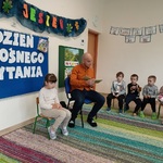 Dzieci siedzą na krzesełkach i słuchają książki czytanej przez mężczyznę.