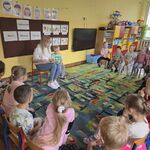 Kobieta trzymająca książkę siedzi na krzesełku naprzeciwko grupy dzieci.