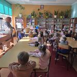 Dzieci siedzą przy stolikach  na krzesełkach w szkolnej czytelni.