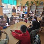 Pani bibliotekarka stoi przed dziećmi_ które siedzą na krzesełkach.