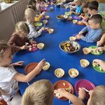 Dzieci siedzą przy połączonych stolikach_ kroją owoce i wkładają do wafelkowych miseczek.