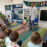 Dzieci siedzą na krzesełkach i patrzą na pana w fartuchu i dziewczynkę trzymającą książkę.