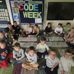 Dzieci siedzą na dywanie wokół napisu code week samodzielnie wykonanego.