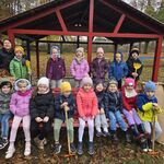  Dzieci zgromadzone są na ławce i wokół niej, niektóre dzieci trzymają grabki, a po obu stronach ławki stoją worki z liśćmi