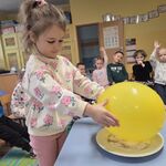 Dziewczynka trzyma żółty balon nad talerzem z pieprzem i cukrem_ w tle siedzi grupa dzieci.