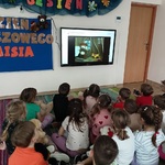 Grupa przedszkolaków siedzi na dywanie i ogląda bajkę.
